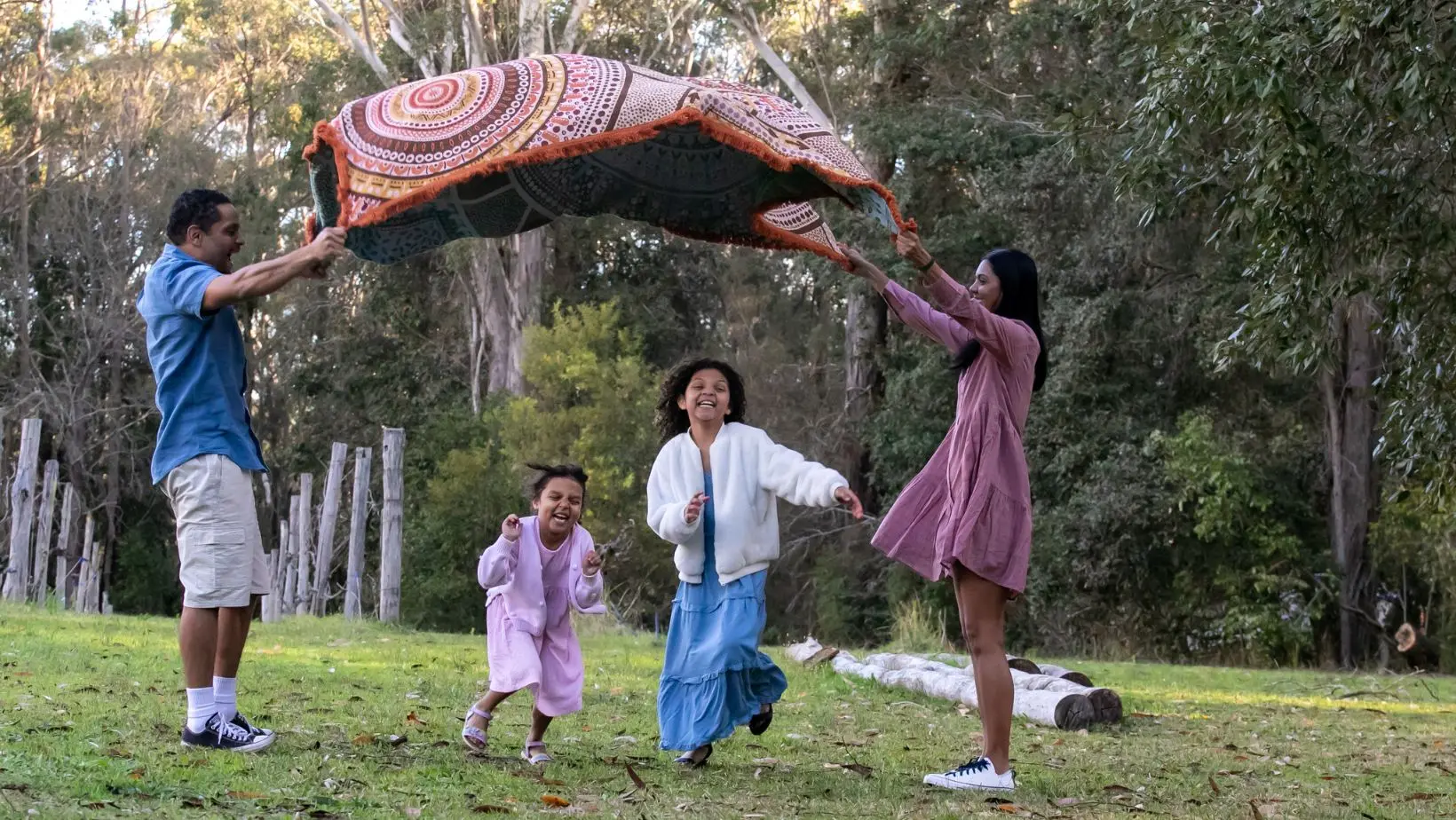 A family laughing outdoors in the bush as two adults lift a patterned blanket over two children